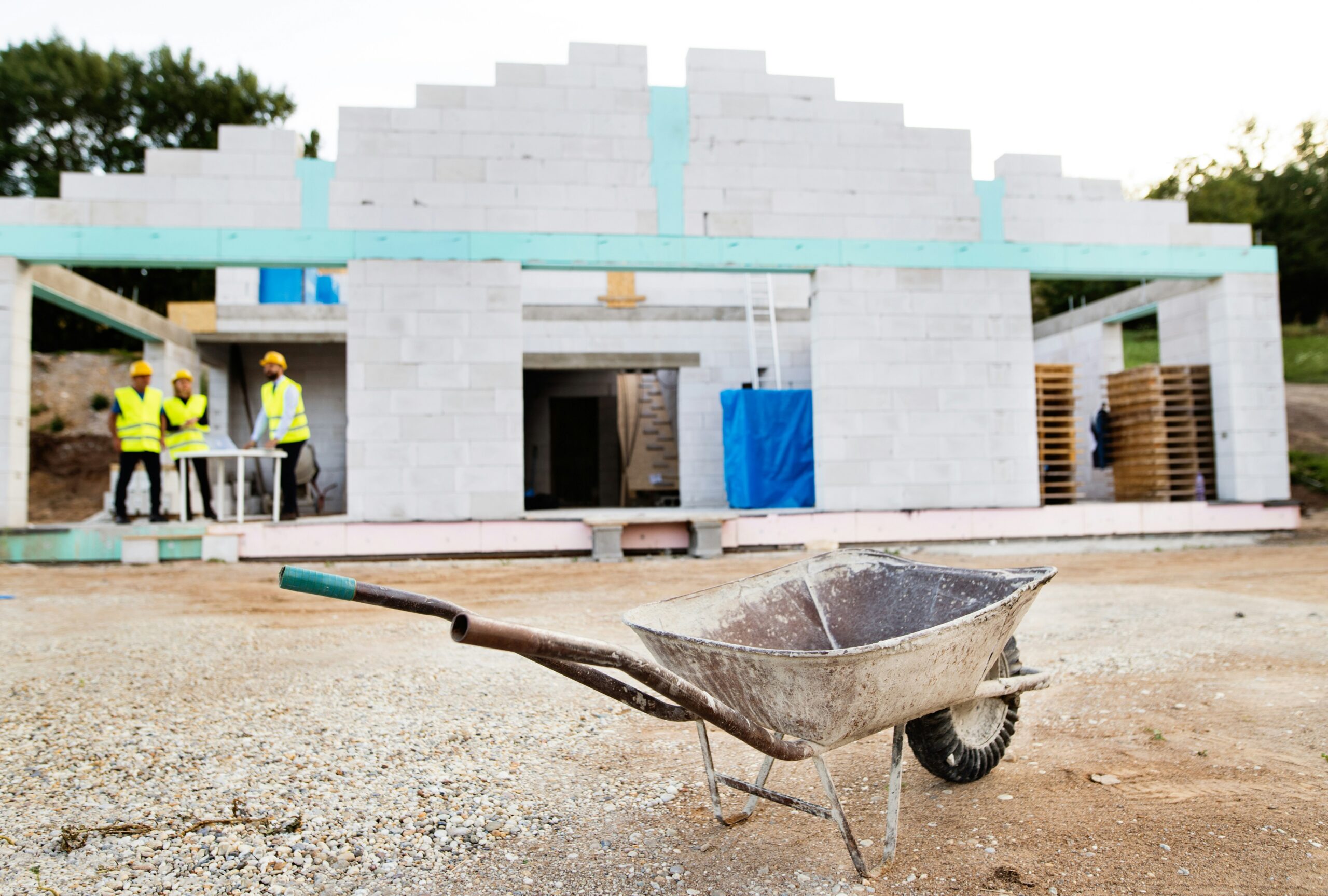 Wheelbarrow in front of a partially constructed concrete block building with construction workers